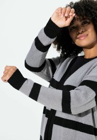 Pitkähihainen harmaa neuletakki, jossa on mustat raidat, resorit hihansuissa ja avoin etuosa. Pehmeä neuletekstuuri ja rento siluetti.