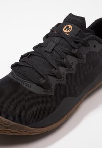 Czarny but sportowy z siateczkową cholewką, płaskimi sznurówkami, szczegółami w formie zygzakowatego przeszycia oraz teksturowaną gumową podeszwą. Logo na języku w kolorze beżowym.