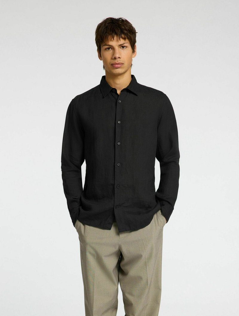 Camisa negra de lino botonada con cuello, mangas largas y un corte relajado, combinada con pantalones beiges.