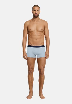 Spierige man die op blote voeten recht vooruit staat, draagt lichtblauwe boxershorts met een donkere tailleband tegen een effen lichte achtergrond.