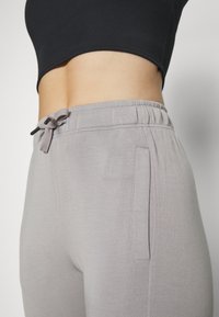 Pantalones de chándal gris claro con cintura ajustable, bolsillos laterales y una textura suave y lisa. La cinturilla presenta un detalle de elástico fruncido.