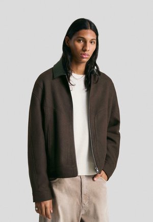 Giacca marrone con zip e colletto verde scuro, design semplice e tessuto texture, indossata sopra un maglione bianco a costine e pantaloni in corduroy beige.