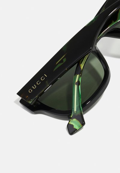 Sončna očala Gucci z bleščečim črnim okvirjem, zelenimi poudarki na ročajih in temnimi lečami. Zlato logotip na vrhu okvirja.