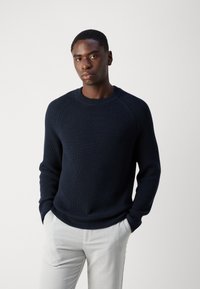 Marineblå ribbestrikket genser med rund hals og lange ermer, kombinert med lyse bukser. Enkel, strukturert design uten tilleggselementer.