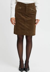 Jupe crayon en velours côtelé marron avec poches zippées, une taille, et une coupe droite, associée à des escarpins noirs à bout pointu et des collants noirs transparents.