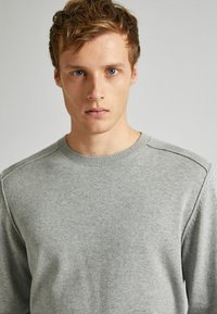 Junger Mann mit kurzem hellbraunem Haar und blauen Augen, der einen schlichten hellgrauen Rundhalsausschnitt-Pullover vor einem neutralen Hintergrund trägt.