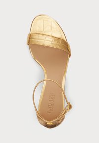 Sandalha de salto dourado com padrão em relevo de croco na parte superior. Apresenta uma fita estreita e fecho no tornozelo, textura suave e design de ponta aberta.