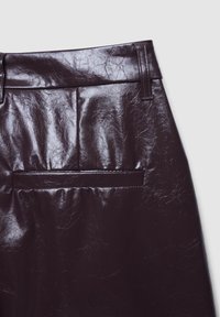 Gros plan d'une jupe en cuir brun foncé brillant avec ceinture, passants et poche passepoilée horizontale arrière sur fond clair.