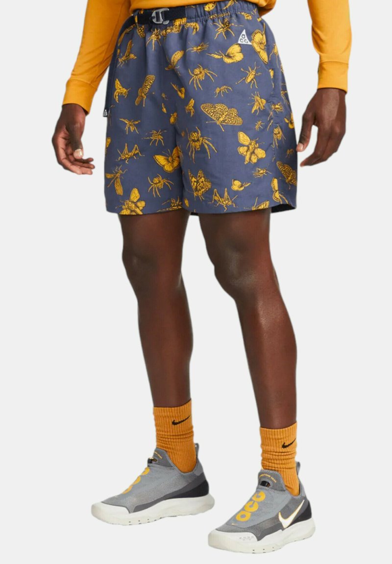 Simshorts i mörkblått tyg med ett mönster av gula insekter. Elastisk midja med justerbar dragsko. (Bärs med grå skor och orange strumpor.)