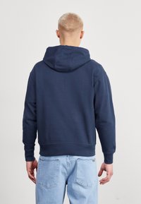 Felpa blu navy realizzata in tessuto morbido, con cappuccio regolabile e polsini a coste. Abbinata a jeans azzurri, vista da dietro.