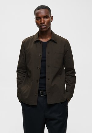 Homme portant une veste boutonnée vert olive foncé par-dessus une chemise noire et un pantalon noir, les mains dans les poches, sur un fond uni.