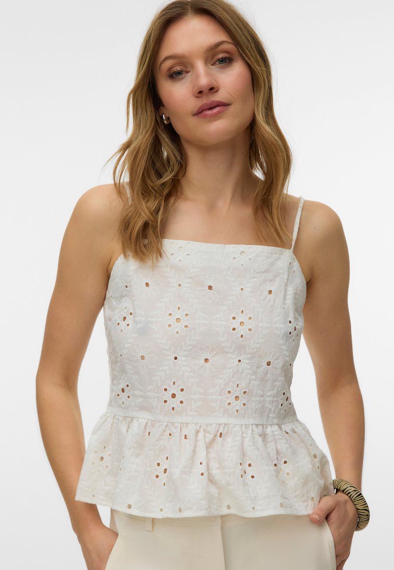 Femme portant un haut sans manches en dentelle à œillets blanc avec un col carré et un ourlet peplum, associé à un pantalon de couleur claire.