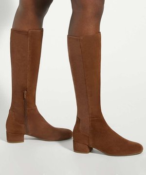 Bottes hautes marron en daim avec des petits talons carrés et des fermetures éclair latérales, portées sur des jambes à peau foncée sur fond blanc.