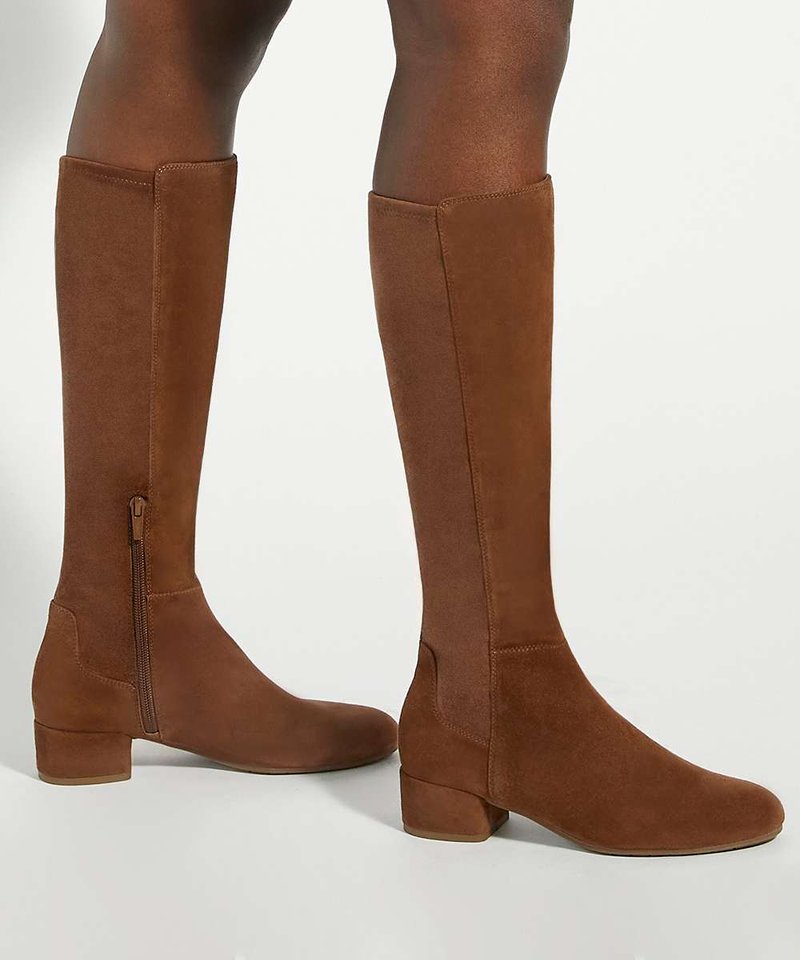 Bottes hautes marron en daim avec des petits talons carrés et des fermetures éclair latérales, portées sur des jambes à peau foncée sur fond blanc.