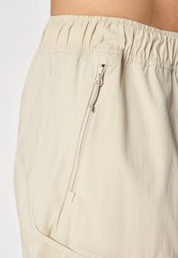 Vaaleanbeige shortsit, jotka on valmistettu sileästä kankaasta ja joissa on joustava vyötärö ja sivussa vetoketjullinen tasku. Yksinkertainen muotoilu, jossa on tasainen siluetti.