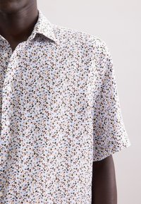 Korte mouwen shirt van licht materiaal, met een klein bloemenpatroon in bruin, groen en blauw op een witte achtergrond. Klassiek kraagontwerp.