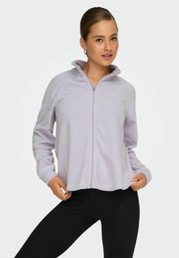 Giovane donna che indossa una giacca in pile viola chiaro con zip e leggings neri, in posa con le mani che tengono i lati della giacca contro uno sfondo bianco.