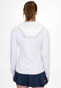 Felpa bianca a maniche lunghe con cappuccio, texture liscia e design aderente, abbinata a una gonna blu navy. La vista di schiena mostra linee pulite.