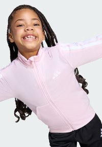 Svijetlo ružičasta jakna sa zatvaračem i visokim ovratnikom, bijelim akcentima na rukavima i prednjim džepovima, u kombinaciji s crnim hlačama. Glatka tekstura.