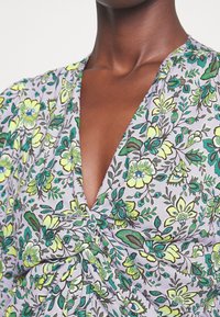 Personne portant une robe à motif floral avec des fleurs vertes et jaunes, avec un profond décolleté en V et un détail de nœud torsadé sur la poitrine.