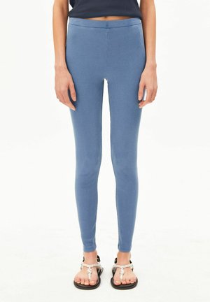 Person trägt eng anliegende blaue gerippte Leggings und schwarze Sandalen mit weißen Riemen, steht vor einem schlichten weißen Hintergrund.