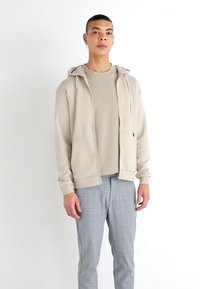 Sudadera beige con cremallera y textura suave, llevada sobre una camiseta de color beige claro, combinada con pantalones de cuadros grises, que tienen un corte ajustado y capucha ajustable.