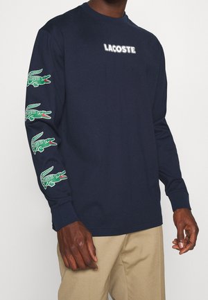 Homme portant une chemise bleu marine à manches longues avec un logo Lacoste blanc sur la poitrine et quatre motifs crocodiles verts sur la manche gauche, associée à un pantalon beige.