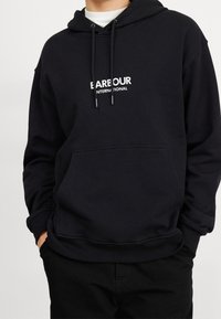 Černá bavlněná mikina s kapucí, která má klokaní kapsu, stahovací šňůrky a bílý nápis „Barbour International“ na hrudi.