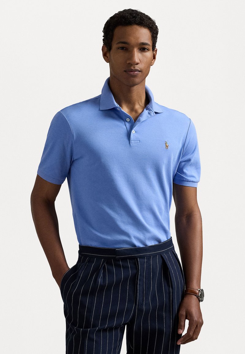 Polo de color azul claro con cuello, tres botones y un pequeño logotipo bordado. Combinado con pantalones oscuros a rayas.