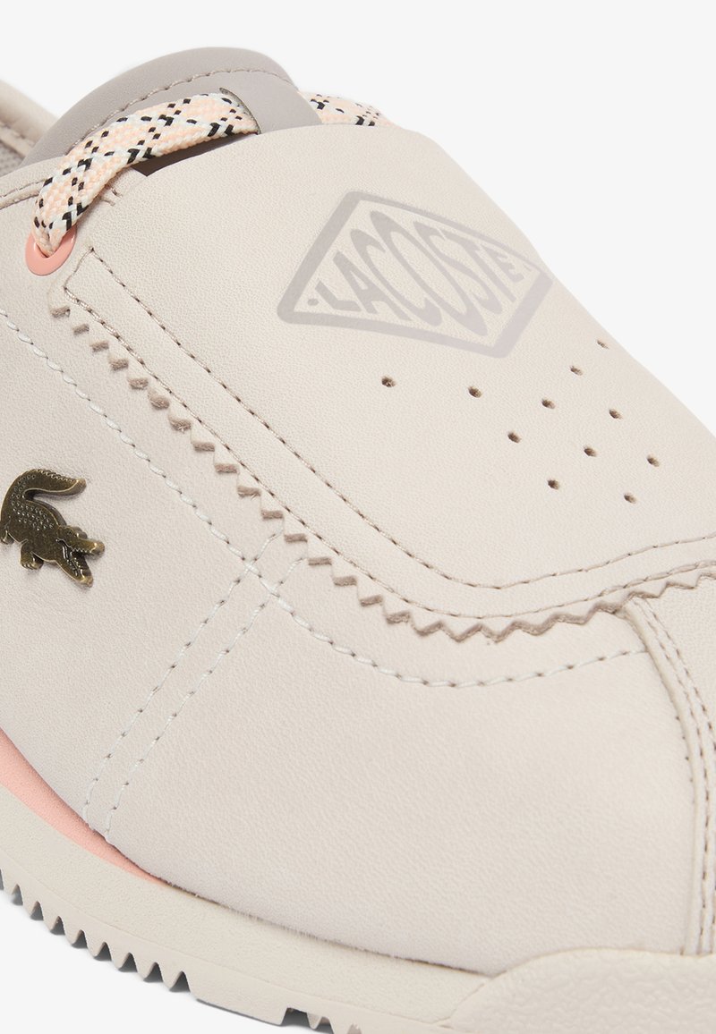 Beiger Lacoste-Sneaker mit hellrosa Akzenten, perforierter Spitze, gemusterten Schnürsenkeln und Metall-Krokodil-Logo an der Seite.