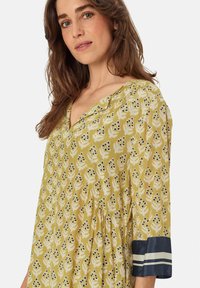 Żółta sukienka w kwiatowy wzór z dekoltem w kształcie litery V, zebranym w pasie oraz granatowymi akcentami na mankietach w paski. Lekki materiał o gładkiej teksturze.