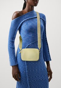 Borsa a tracolla gialla in pelle con una texture a buccia d'arancia e chiusura con zip. Presenta una fascia ampia e regolabile sopra un vestito blu a costine.