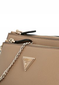 Borsa Guess beige texturizzata con logo triangolare dorato, tracolla in catena argentata e zip dorate su sfondo bianco.