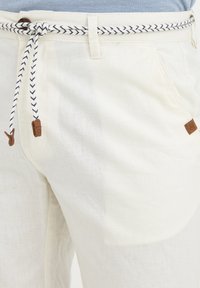 Hvit linbukser med en flettet marineblå og hvit snor, to sidelommer, og brune lærdetaljer med logo.