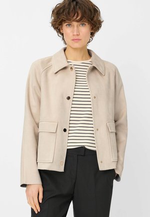 Frau mit kurzen lockigen Haaren, die eine beige, geknöpfte Jacke über einem schwarz-weiß gestreiften Shirt und schwarzen Hosen vor einem einfarbigen Hintergrund trägt.