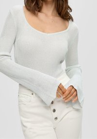 Maglione a coste di un blu chiaro con scollatura profonda e maniche lunghe svasate, abbinato a pantaloni bianchi a vita alta con dettagli decorativi sui bottoni.