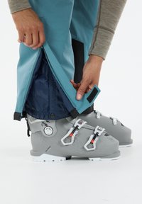 Skidstövlar i ljusgrått med justerbara spännen, i kombination med blå skidbyxor. Byxorna har en dragkedja och elastisk manchett för anpassning.