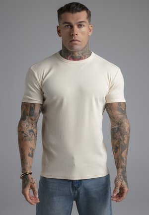 Homme aux cheveux courts, tatouages sur le cou et le bras, portant une chemise beige à manches courtes et un jean bleu, debout devant un fond gris uni.