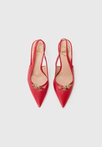 Zapatos de tacón de piel roja con punta afilada, que presentan una hebilla decorativa en forma de corazón dorado y detalles recortados en los lados.
