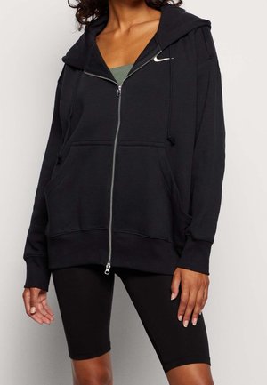 Femme portant un sweat à capuche zippé noir avec un logo Nike blanc et un short de cyclisme noir, debout les mains détendues le long du corps.