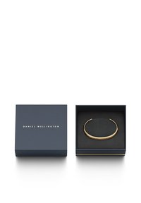 Gold cuff bracelet displayed in a navy blue box. Smooth metal surface with a slight curve, featuring engraved brand name. Black velvet lining.