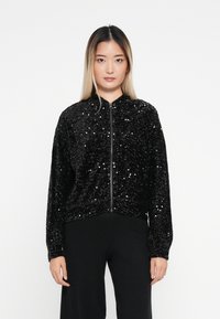 Czarna bomberka z cekinami, zapinana na zamek, z prążkowanymi mankietami i dołem, posiadająca miękką teksturę i luźny krój. Nosi ją z czarnymi spodniami.