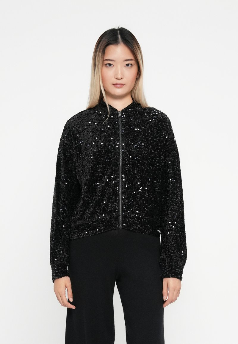 Czarna bomberka z cekinami, zapinana na zamek, z prążkowanymi mankietami i dołem, posiadająca miękką teksturę i luźny krój. Nosi ją z czarnymi spodniami.