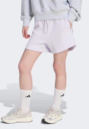 Osoba ubrana w jasnofioletowe szorty, białe skarpetki sportowe z czarnym logo, jasnoszare sneakersy i szarą bluzę z rękami w kieszeniach.
