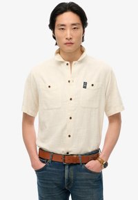 Camicia a maniche corte con colletto di un beige chiaro, dotata di due tasche sul petto e bottoni marroni. Indossata con jeans in denim blu e una cintura marrone.