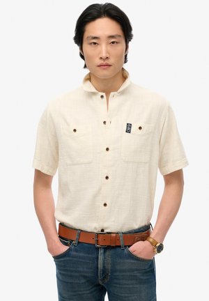 Camisa de manga corta y cuello en beige claro, con dos bolsillos en el pecho y botones marrones. Llevada con jeans de mezclilla azules y un cinturón marrón.