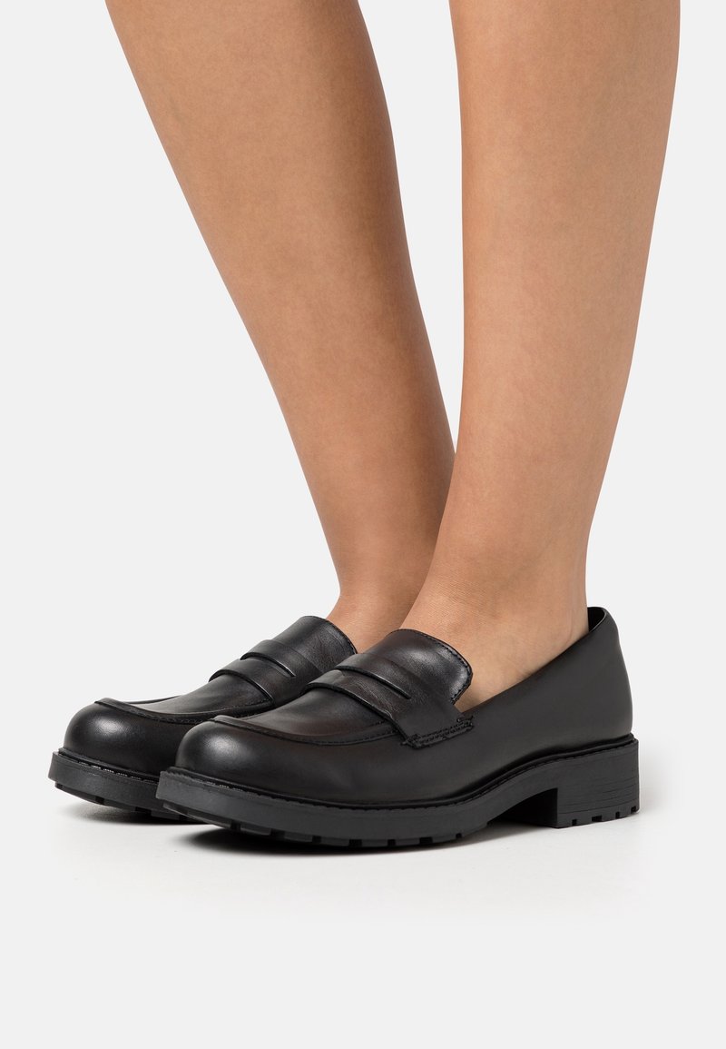Mocassins en cuir noir à semelles épaisses portés pieds nus, vus de côté avec les jambes visibles jusqu'à mi-mollet sur un fond neutre.