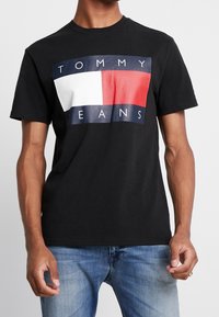 Černé tričko s krátkým rukávem, které má velký logo design v červené, bílé a modré barvě s textem "TOMMY JEANS" ve spodní části. Klasický styl s kulatým výstřihem.