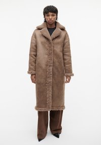 Bruine nepleren shearlingjas met een grote kraag, voorknoopsluiting en getextureerde afwerking. Model draagt brede bruine broeken en puntige zwarte hakken.