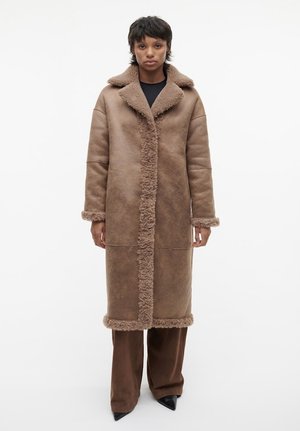 Bruine nepleren shearlingjas met een grote kraag, voorknoopsluiting en getextureerde afwerking. Model draagt brede bruine broeken en puntige zwarte hakken.